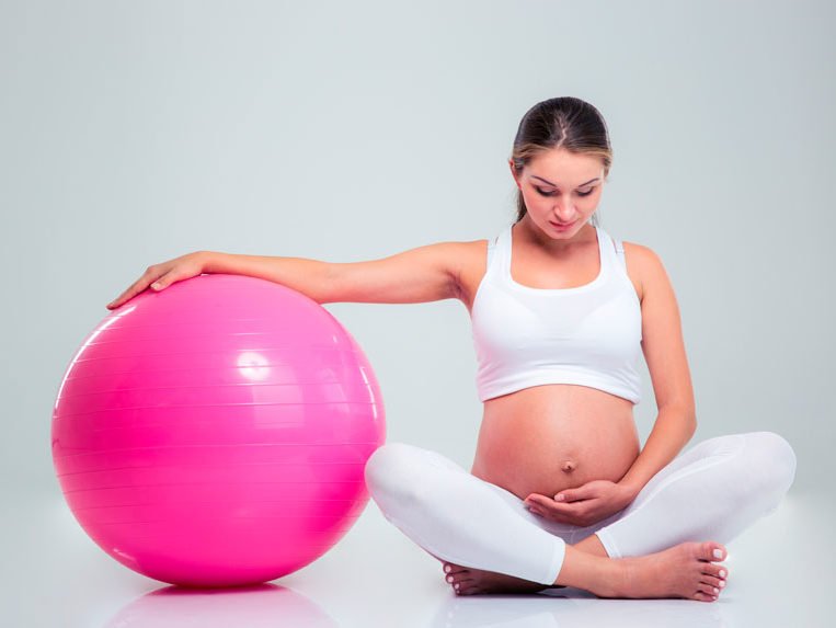 Jovem grávida apoia um braço numa bola de pilates grande, cor de rosa