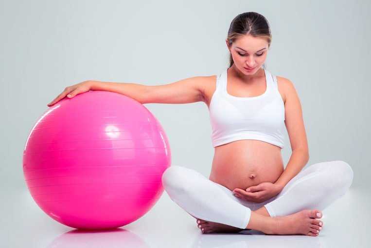 Jovem grávida apoia um braço numa bola de pilates grande, cor de rosa